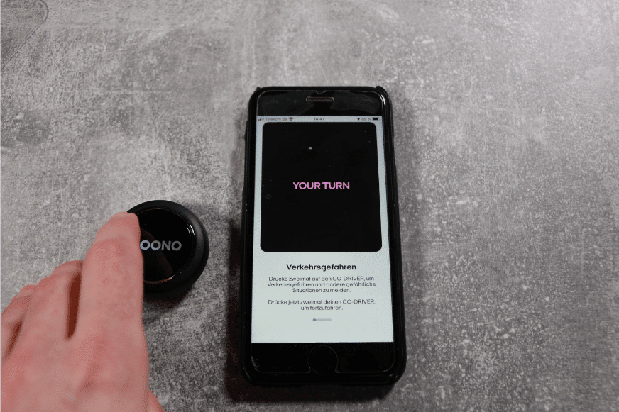Der Ooono Co-Driver No1 liegt neben dem Smartphone, wird gedrückt und es wird erklärt wie Du andere vor Verkehrsgefahren warnen kannst